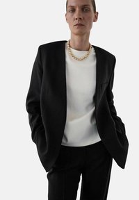 Zwarte tailor-made blazer met scherpe schouders, witte top eronder en een chunky gouden kralenketting. Gladde stof, minimalistisch ontwerp.