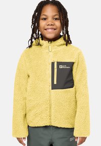 Keltainen fleece-takki, jossa on teksturoidun pinnan viimeistely, vetoketjukiinnitys ja tummanharmaa rintatasku, jossa on logo. Vaatteena oli oliivinvihreät housut.