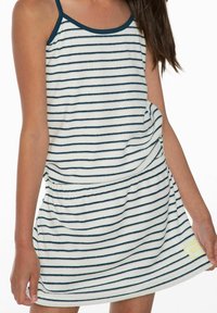 Dziewczyna w bezrękawowej białej sukience w cienkie poziome granatowe paski i granatowym dekoltem, trzymająca dół sukienki obiema rękami.