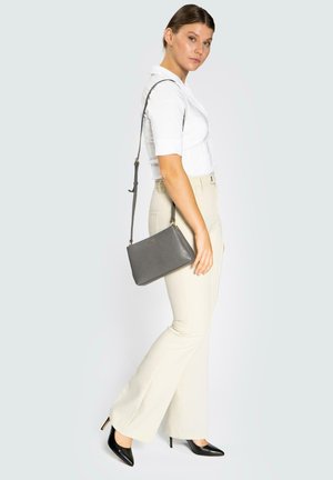 Sac en bandoulière en cuir gris avec une surface texturée, une quincaillerie dorée, une forme rectangulaire et une sangle réglable, porté avec un pantalon évasé beige.