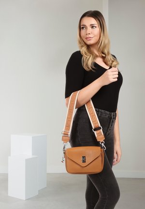 Vrouw draagt een zwarte top en donkere jeans, met een tan leren handtas met brede, patroonband over haar schouder, tegen een effen muur.