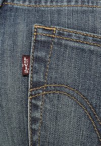 Kangas tummaa sinistä farkkua, jossa näkyy taskun sauma. Pienessä punaisessa Levi's-tarrassa on kiinnitys, ja kultaiset ompeleet korostavat taskua.