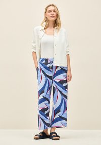 Losse, patroonrijke broek in blauw met rode en marineblauwe bladdessins, gecombineerd met een witte overhemd met knopen en donkere sandalen.