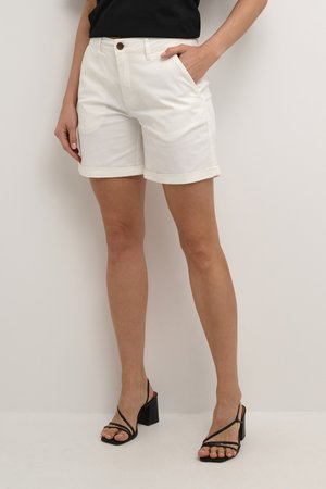 Shorts en coton blancs avec des revers, poches avant et fermeture à bouton en métal, accompagnés de sandales à talons noires à lanières.