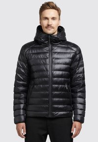 Bărbat îmbrăcat cu o jachetă puffer neagră cu umplutură și glugă, stând cu fața spre camera pe un fundal simplu deschis.