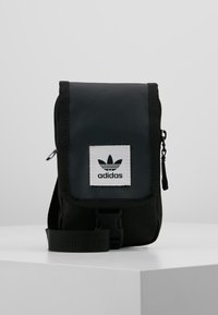 Bolso bandolera negro de nailon con tapa de cierre, cuenta con un parche del logo de Adidas en blanco, correa ajustable y bolsillo delantero con cremallera.