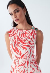 Sarkani un balta kleita bez piedurknēm ar abstraktu rakstu, teksturētu audumu un dekoratīvu plecu akcentu ar krāsainām pērlītēm.