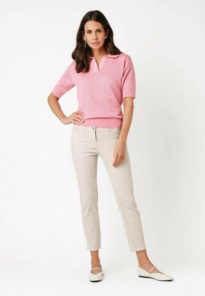 Woman with long dark hair wearing a pink short-sleeve knit top, beige pants, white flats, and silver earrings, standing with one hand on hip.