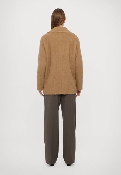 Brązowy puszysty płaszcz z kołnierzem, opadającymi ramionami i luźnym krojem. Połączony z szarymi spodniami o szerokich nogawkach.