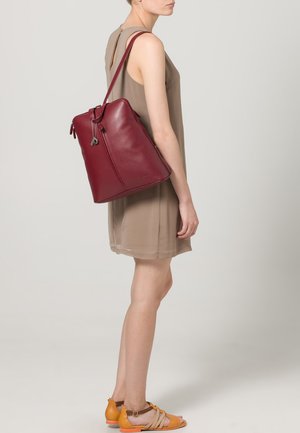 Sac à dos en cuir rouge, de forme rectangulaire avec fermeture éclair, avec une bandoulière réglable et un accent de porte-clés, porté avec une simple robe beige.