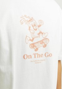 Witte katoen T-shirt met een lichtroze grafiek van een cartoon die aan het skateboarden is, met de tekst "On The Go" eronder.