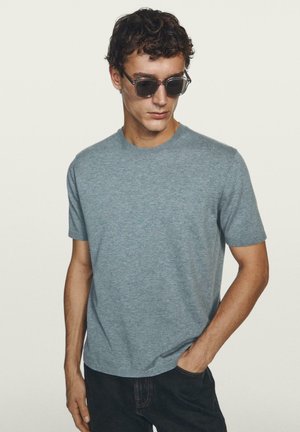 Uomo con indosso una maglietta grigia a maniche corte e occhiali da sole, con una mano in tasca, che guarda verso il basso su uno sfondo chiaro e uniforme.