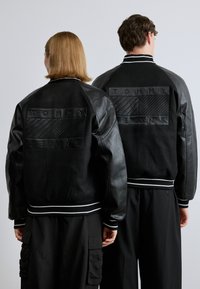 Jachetele tip bomber din piele neagră prezintă panouri texturate, manșete și tiv striate în alb contrastant și logo-ul „TOMMY JEANS” în relief pe spate.