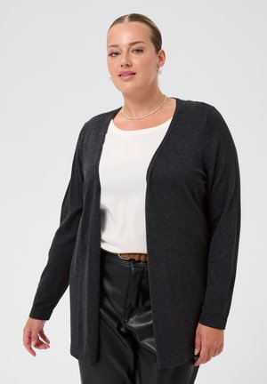 Femme portant un cardigan gris foncé ouvert devant sur un haut blanc avec un pantalon en cuir noir et une ceinture marron, debout devant un fond uni.