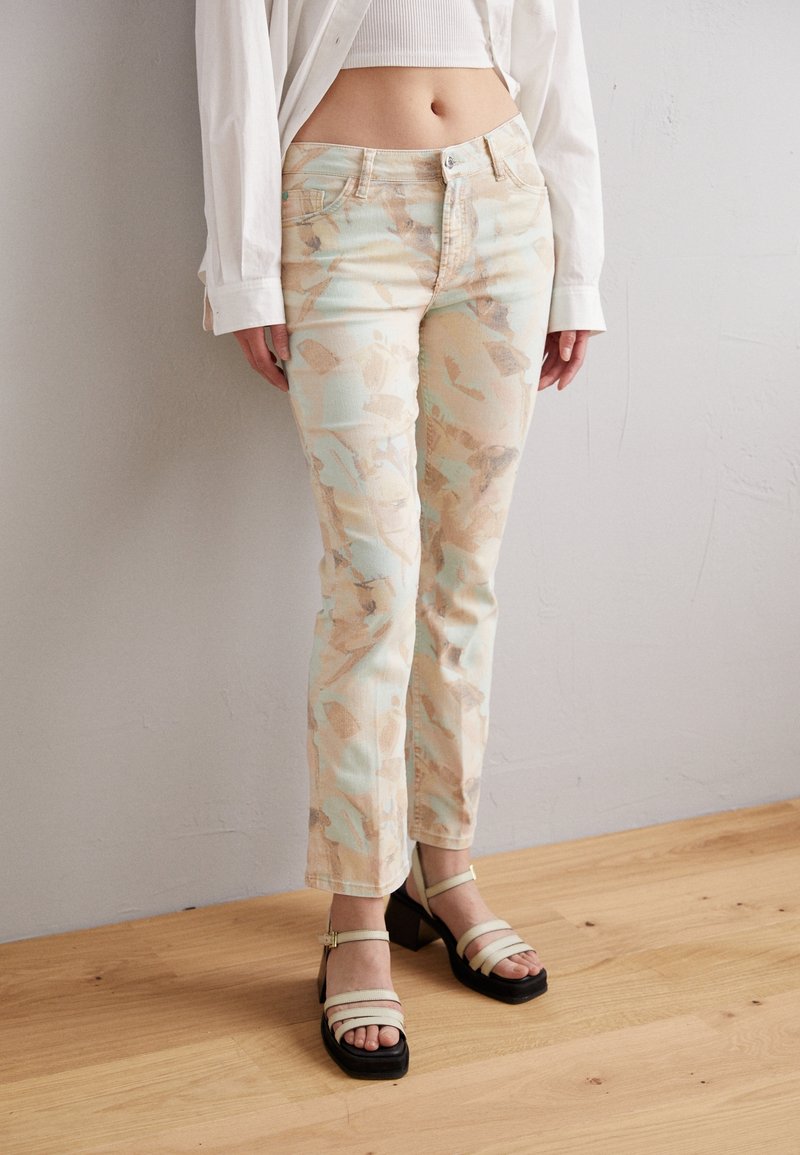 Femme portant un pantalon clair à motifs, un top court blanc, une chemise blanche à manches longues et des sandales beiges à talons, debout sur un sol en bois.