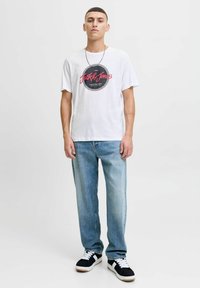 Witte T-shirt met korte mouwen en ronde grafische print; lichtblauwe relaxed-fit jeans; zwart-witte sneakers met gumzolen; zilveren ketting.