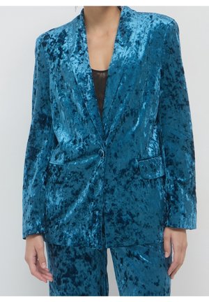 Femme portant un blazer en velours froissé brillant couleur bleu sarcelle avec un pantalon assorti et un haut noir transparent en dessous.