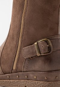 Brązowy zamszowy i skórzany but z bocznym suwakiem, regulowanym paskiem z mosiężną klamrą oraz ćwiekowanym wykończeniem powyżej teksturowanej podeszwy.