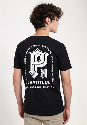 Ung mand med krøllet hår iført en sort t-shirt med hvidt grafisk tekst om taknemmelighed og Bolingbrook, Illinois på ryggen.