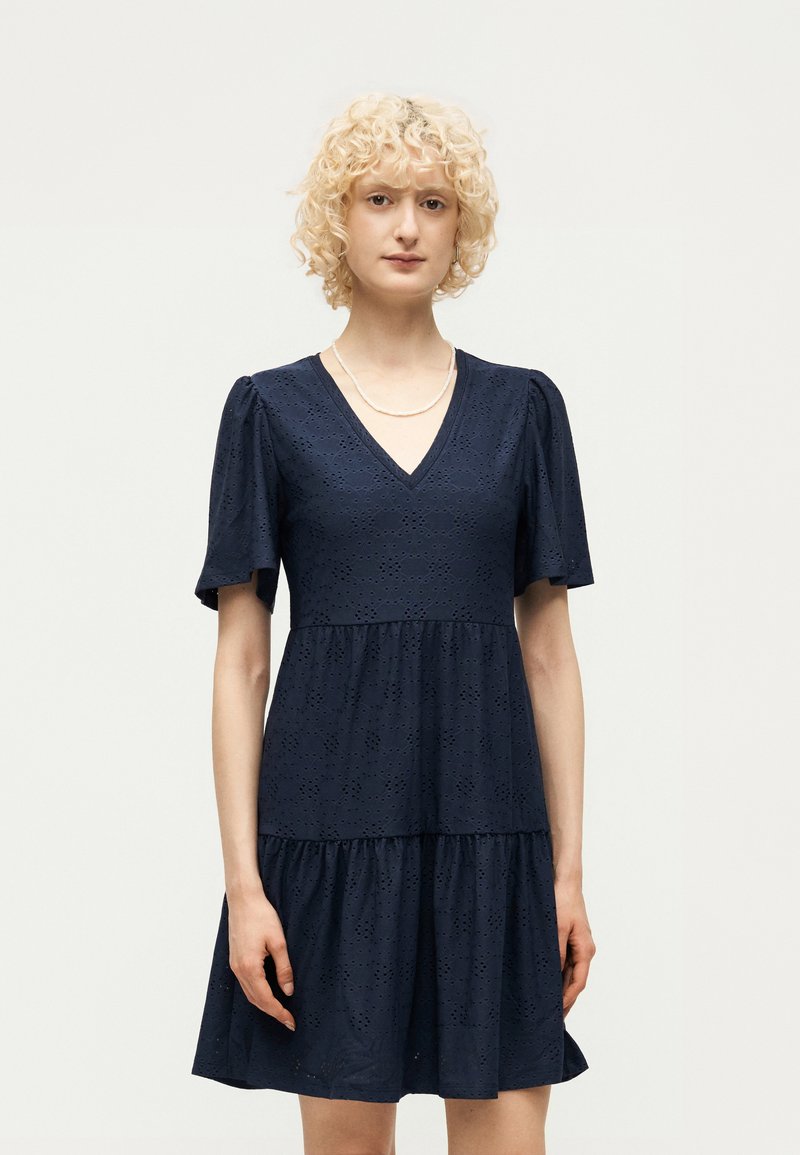 Νεαρό άτομο με σγουρά ξανθά μαλλιά που φοράει φορέμα navy μπλε με τρυπητά σχέδια, κοντά μανίκια και V-λαιμόκοψη, στέκεται μπροστά σε απλό φωτεινό φόντο.