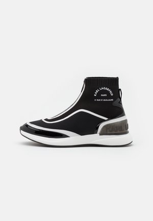 Chaussure noire en maille tissée à la cheville avec des accents blancs, un bout brillant, un logo à l'arrière et une semelle blanche épaisse. Dotée d'un design sans lacets et d'un matériau flexible.