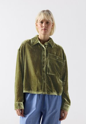 Blonde Person trägt ein locker sitzendes, olivgrünes Samthemd mit Knöpfen und großer Brusttasche sowie blaue gestreifte Hose vor einfachem weißem Hintergrund.