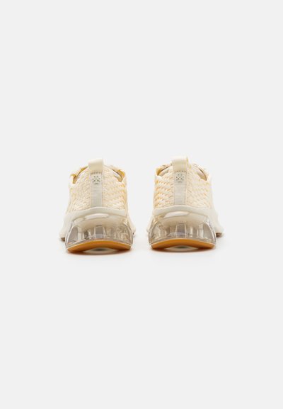 Sneakers beige chiaro con una tomaia in maglia strutturata, suola trasparente e dettagli in gomma. Presentano un passante sul retro per facilitarne l'indossamento.