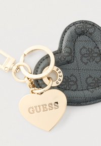 Sort tekstureret læderhjerte-nøglering med guldfarvet hardware og "GUESS"-logo på en metallabel, med to fastgjorte metalringe.