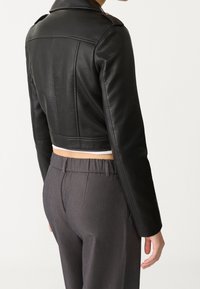 Veste en cuir noire courte présentant une texture lisse et un design structuré. Associée à un pantalon taille haute gris avec une taille froncée.