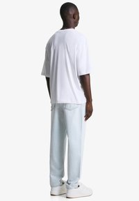 Bijela prevelika majica s kratkim rukavima, u kombinaciji s svijetloplavim trapericama ravnog kroja i bijelim tenisicama, viđena sa stražnje strane.