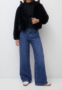 Czarna kurtka z sztucznego futra, krótka, z kapturem, w połączeniu z luźnymi niebieskimi dżinsami i białymi trampkami z jasnobrązową podeszwą. Widoczny zamek.