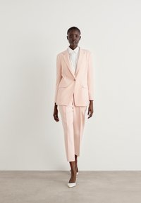 Helles rosa maßgeschneidertes Anzug mit einem taillierten Blazer und geraden Hosen, kombiniert mit einem weißen Hemd mit Kragen und weißen Schuhen mit spitzem Zeh.