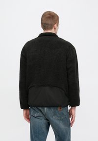 Giacca in pile nera con una parte superiore testurizzata e soffice, caratterizzata da una sezione inferiore liscia nera e un colletto. Indossata sopra dei jeans blu.