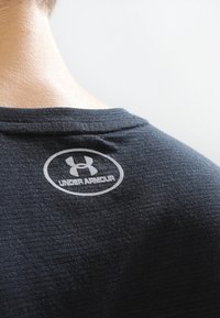 Black athletic shirt featuring a round neck and textured fabric. Displays a reflective Under Armour logo on the back near the collar.
