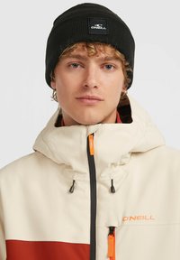 Beige og rustfarvet farveblokeret vandtæt jakke med hætte, sort lynlås og orange detaljer. Båret med en sort ribbet hue med et logo-patch.