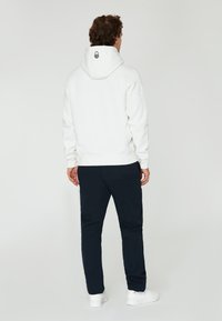 Sudadera blanca con cordón y logo negro en la parte posterior, combinada con pantalones azul marino oscuros y zapatillas blancas, mostrando un ajuste relajado.
