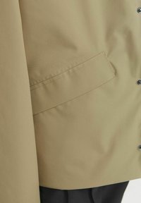 Strak, beige buitenkleding met een gladde textuur, voorzien van een klepzak, drukknoopsluiting en een ontspannen pasvorm, gedeeltelijk over een zwart kledingstuk gedrapeerd.