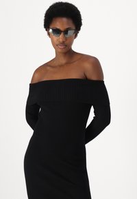 Abito nero a coste scollato sulle spalle con maniche lunghe; texture liscia; design aderente. Modella con occhiali da sole scuri e angolari.