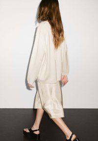 Maglione beige oversize, gonna satin al ginocchio in beige chiaro e sandali con platform neri. Tessuti morbidi, vestibilità rilassata, design minimalista.