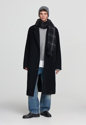 Giovane uomo che indossa un berretto grigio, un soprabito nero, un maglione grigio, una sciarpa a quadri, jeans larghi blu e scarpe di pelle nere, in piedi su uno sfondo semplice.