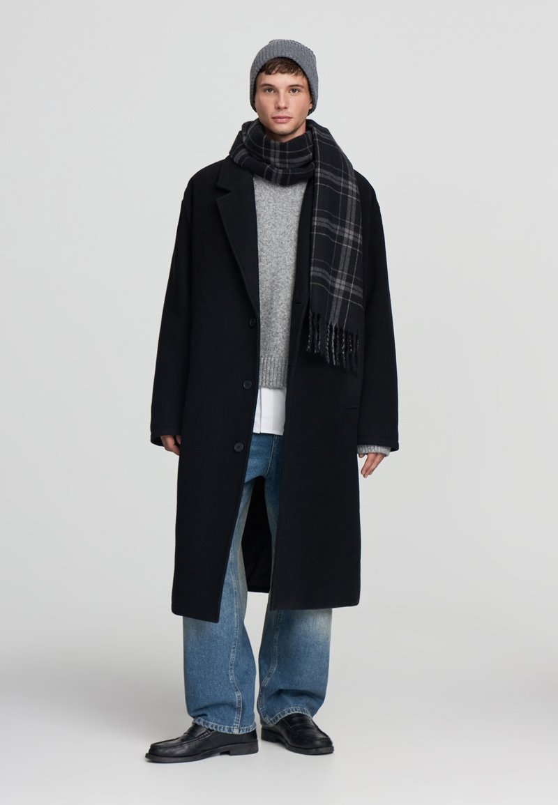 Giovane uomo che indossa un berretto grigio, un soprabito nero, un maglione grigio, una sciarpa a quadri, jeans larghi blu e scarpe di pelle nere, in piedi su uno sfondo semplice.