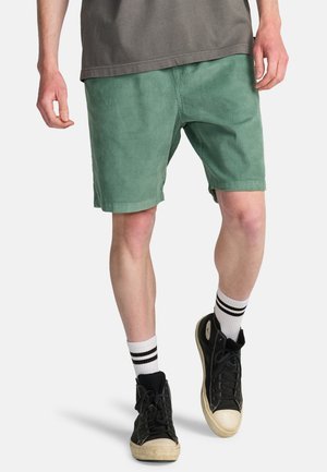 Pantaloncini verdi in velluto a coste con una vestibilità rilassata, tasche laterali e una texture liscia. Abbinati a sneaker nere e calzini bianchi a righe.