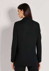 Frau mit langen, dunklen Haaren, die ein schwarzes Blazer und auffällige, hängende Ohrringe trägt, von hinten gegen einen schlichten, hellen Hintergrund betrachtet.