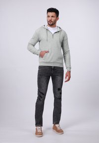 Man wearing light gray zip-up hoodie, white t-shirt, black ripped jeans, and brown sneakers standing with one hand in pocket.