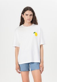 Camiseta blanca de algodón con un limón amarillo y una hoja verde bordados en el lado izquierdo del pecho, combinada con pantalones cortos de mezclilla.