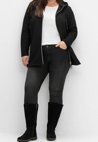 Donna in giacca nera decorata, maglietta bianca tipo henley, jeans scuri e stivali neri scamosciati fino al polpaccio, in piedi su uno sfondo bianco.