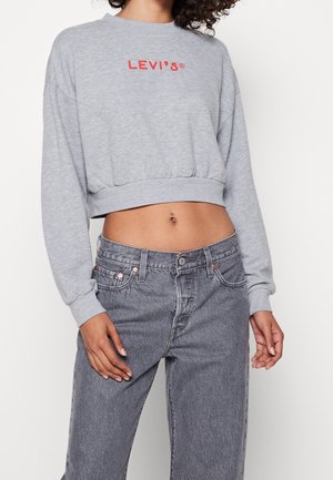 Kvinde iført grå kort Levi's sweatshirt og højtaljede, falmede sorte jeans, der viser en navlepiercing, stående foran en ensfarvet baggrund.