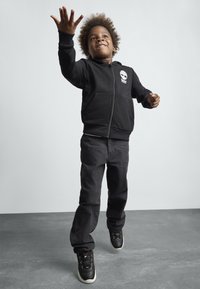 Svart hettegenser med glidelås, frontlommer og logo, sammen med mørke bukser og svarte joggesko. Barnet er i en lekfull posisjon med armene utstrakt.