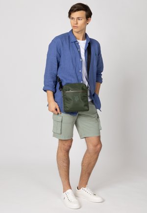 Borsa a tracolla verde in nylon con chiusura lampo, logo ricamato e tracolla regolabile, indossata su una camicia blu e pantaloni cargo verdi.