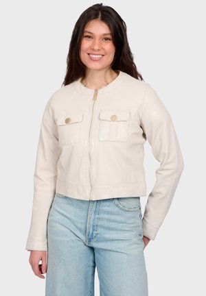 Young woman smiling, wearing a light beige zip-front jacket with buttoned chest pockets and light blue high-waist jeans.